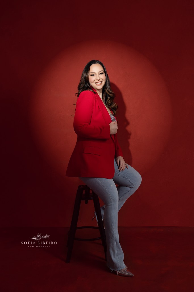 woman poses in a blazer and jeans combo against a red backdrop for birthday photos
