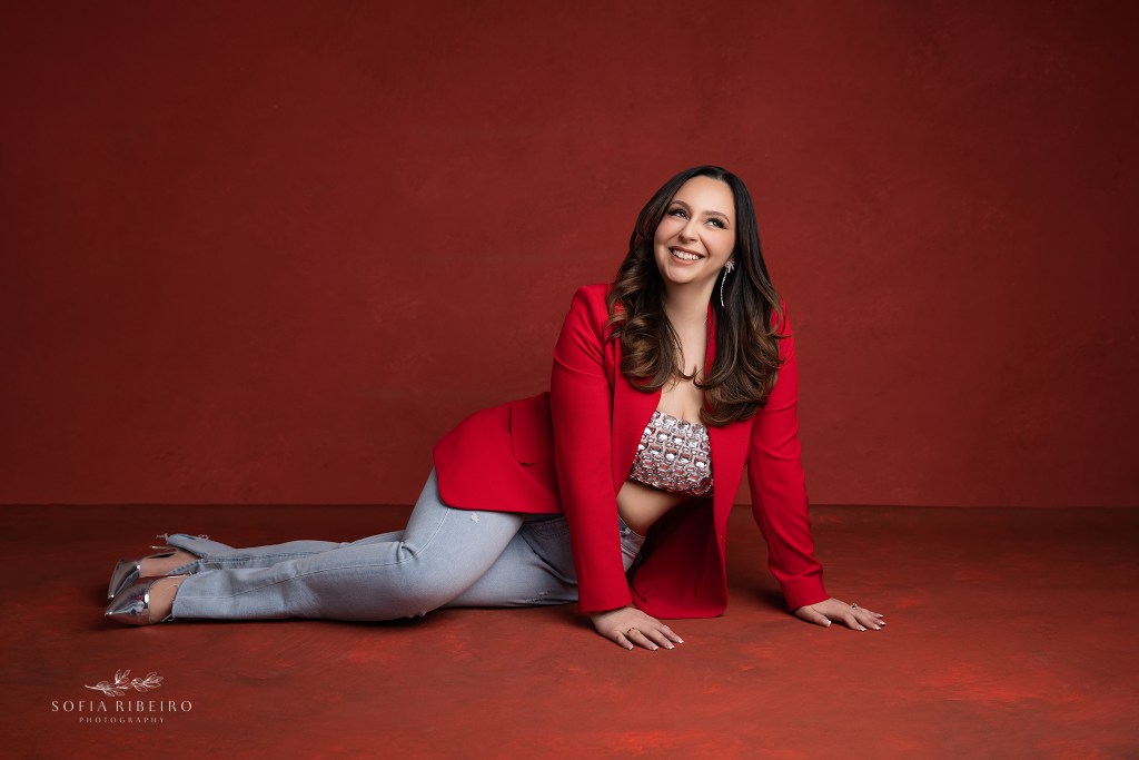 woman poses in a blazer and jeans combo against a red backdrop for birthday photos
