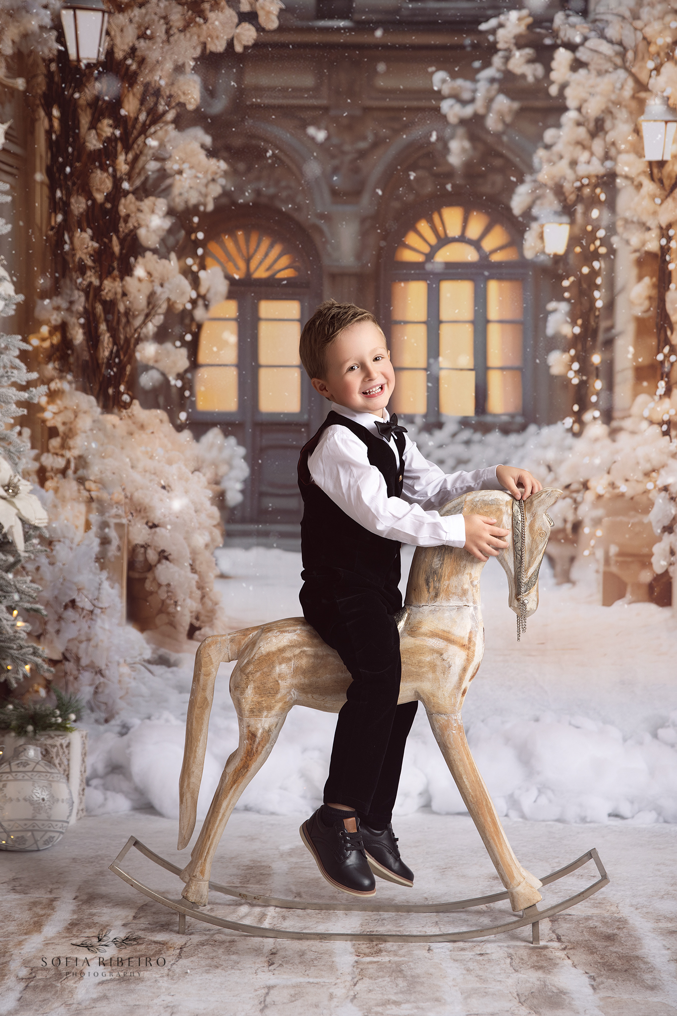 A happy young boy in a black vest and slacks rides an old wooden rocking horse in the snow