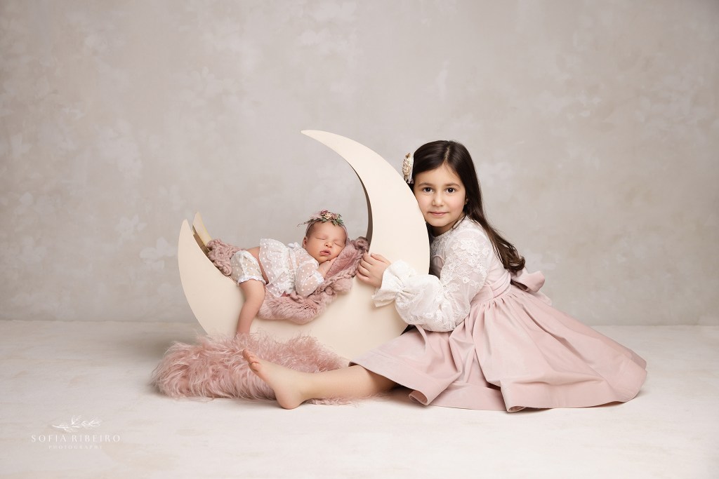 dunellen nj newborn photographer poses a newborn sweetly sleeping in a moon prop with her older sister snuggling the moon and softly smiling