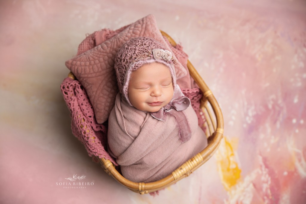 baby girl is swaddled in pink, snuggled in a tiny basket against a pink backdrop