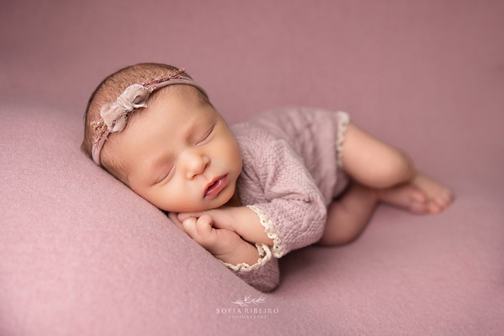 a baby sleeps softly with her hands cradled under her face, dressed in a pink outfit for a newborn photo