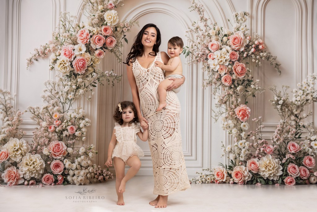 mom poses with her two babies in shades of cream against a floral arch backdrop for mommy and me photos in nj