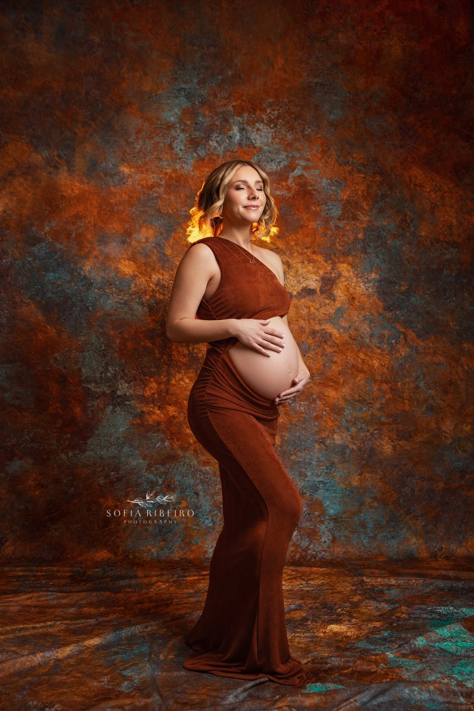 mom to be poses against a rust textured backdrop with a matching body con dress with bump exposed in a weehawken nj maternity photo studio