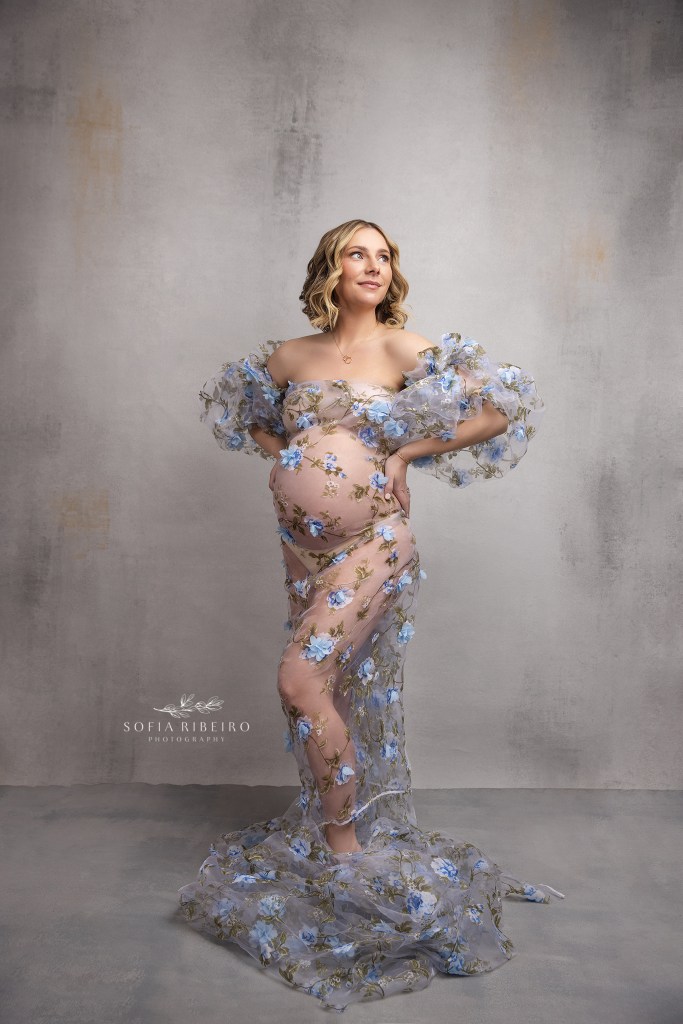 mom to be poses gracefully in a sheer floral applique fabric against a grey backdrop for photos with weehawken nj maternity photographer