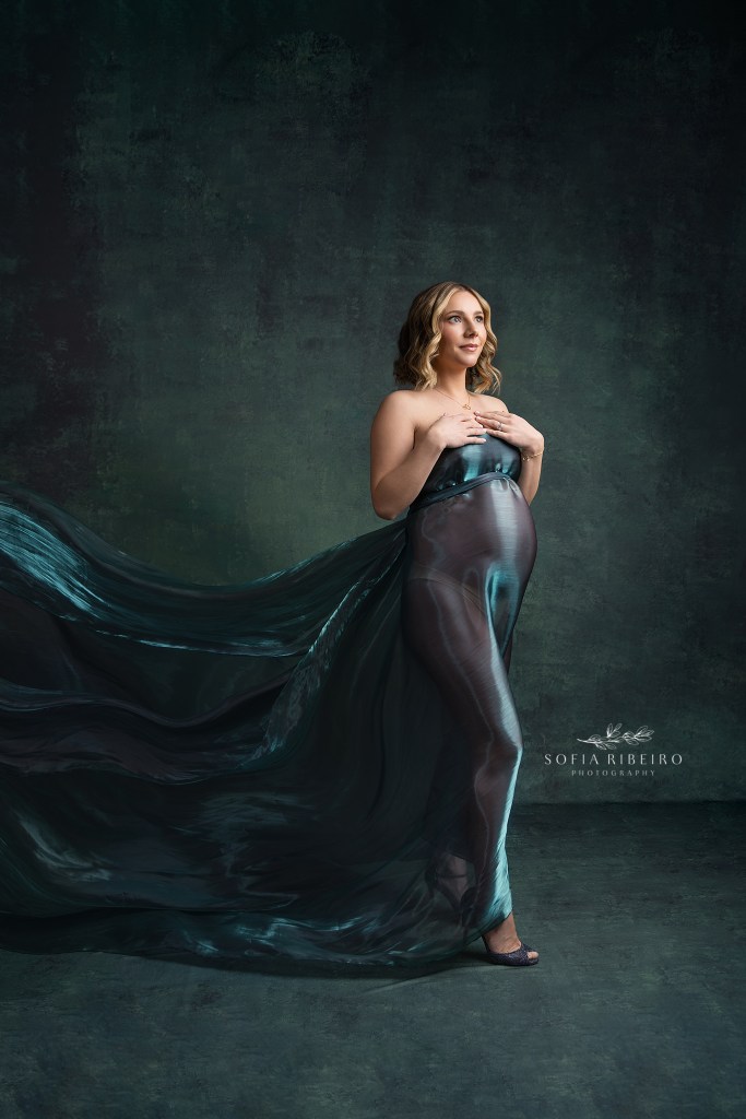 mom is draped in a shimmering teal semi sheer fabric with light falling on her baby bump with fabric flowing behind her