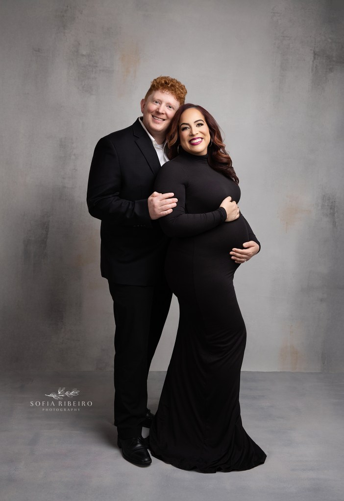 mom and dad smile to camera while showing off the baby bump, dressed in matching b lack outfits