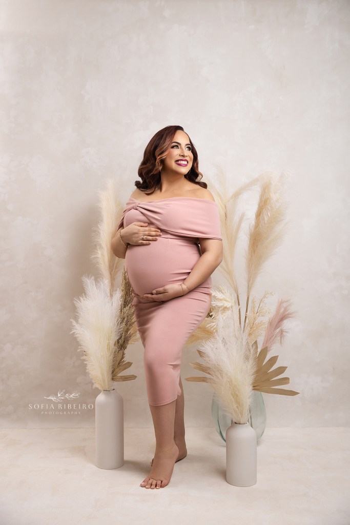 mom poses with beautiful light on her face, cradling her baby bump against a cream backdrop with pampas grasses