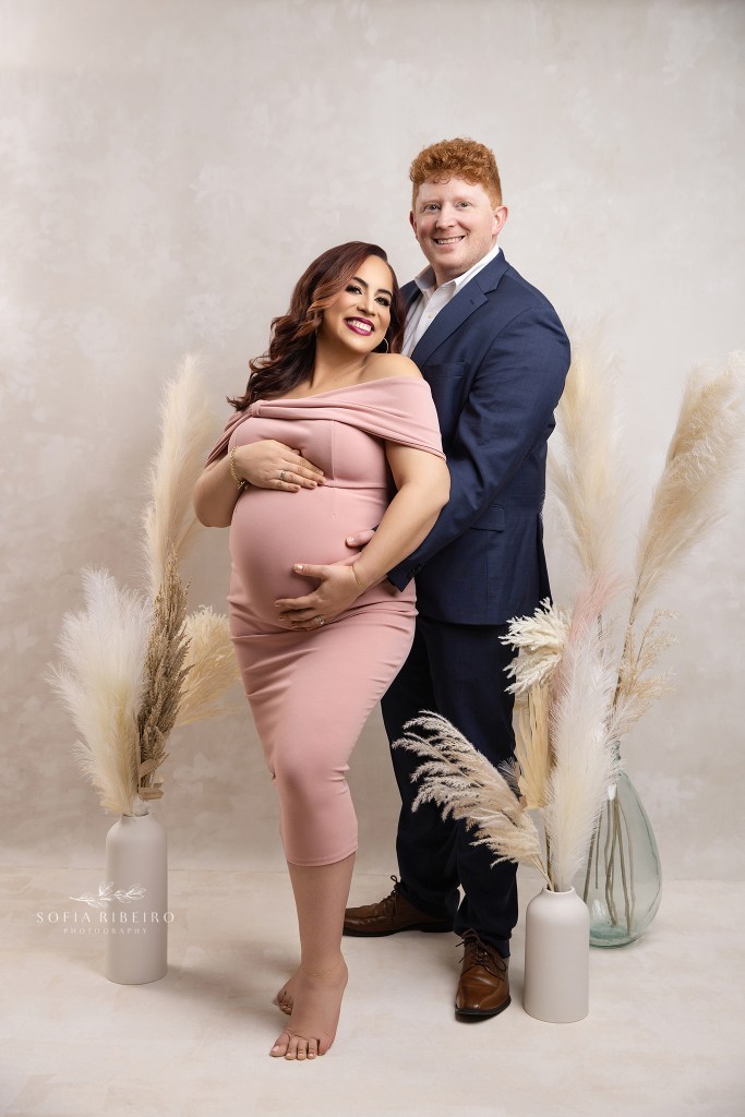 mom and dad pose proudly together in formal outfits, with hands on baby bump for a portrait in a whippany nj photography studio