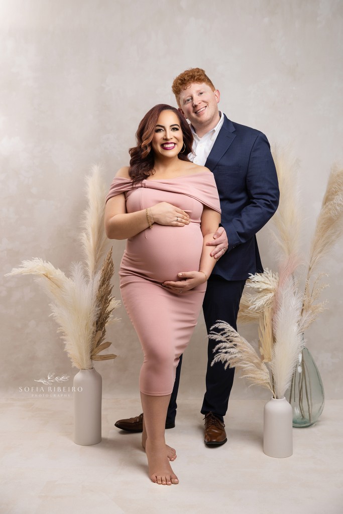 mom and dad pose proudly together in formal outfits, with hands on baby bump for a portrait in a whippany nj photography studio