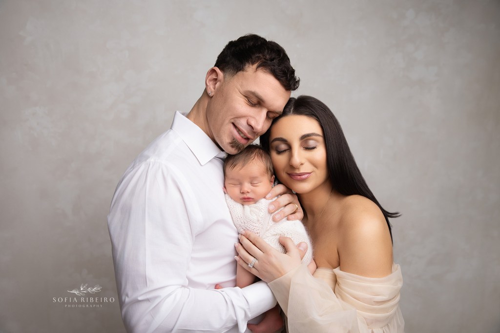 parents smile sweetly together with their new baby against a neutral backdrop during a newborn portrait session