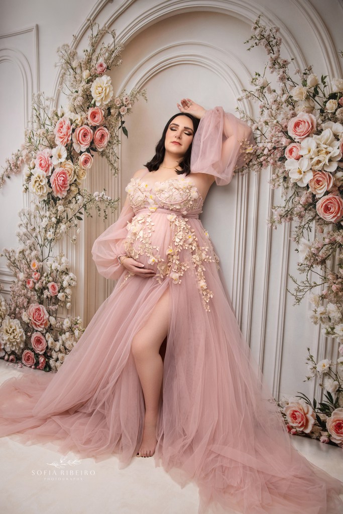 expectant mom in a pink dress leans against a floral backdrop in hudson cally ny