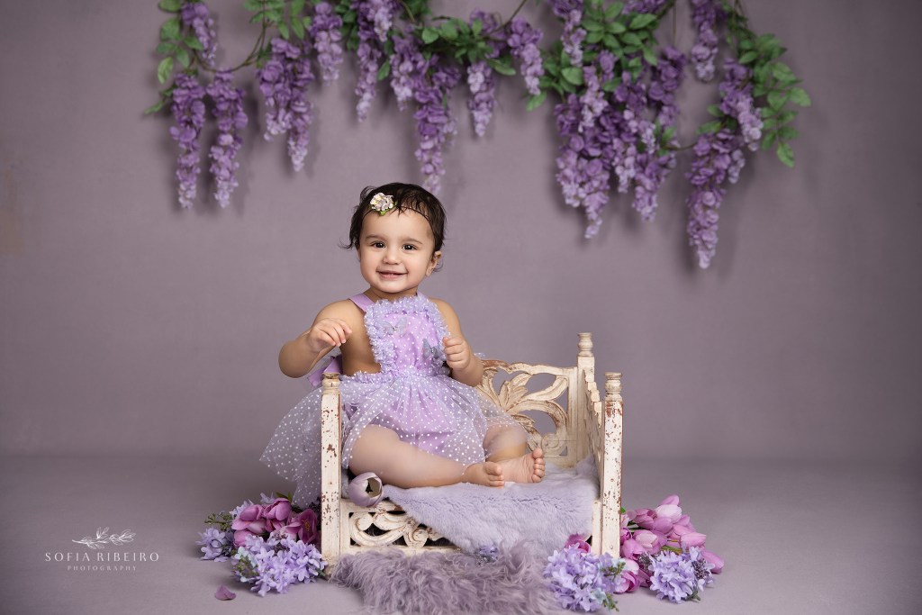 a one year old baby girl poses against a purple backdrop in a sweet dress and bed prop