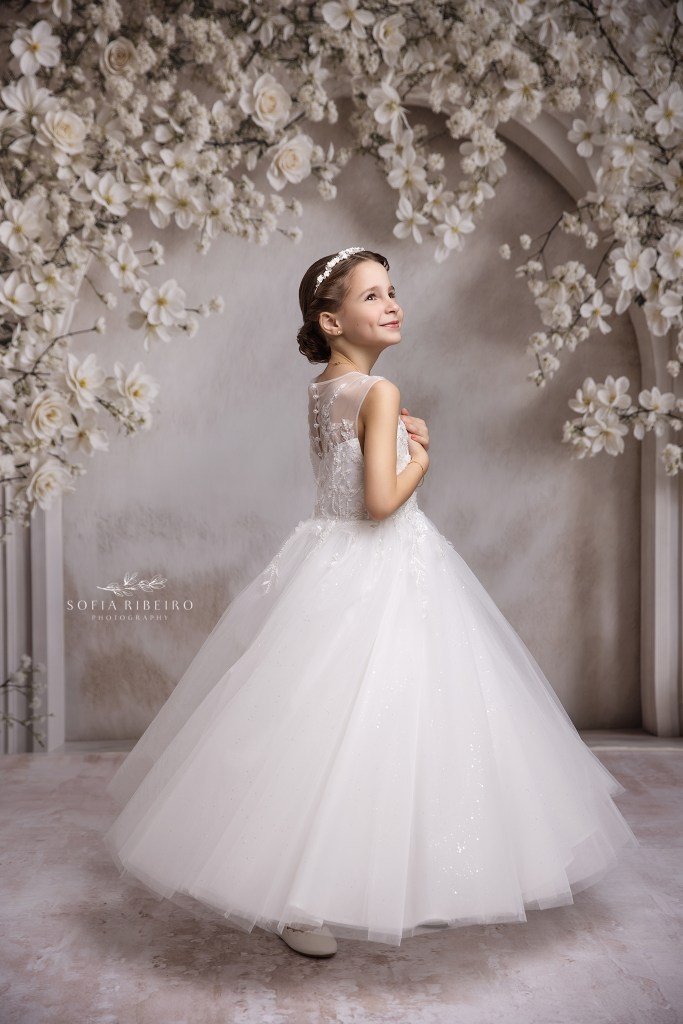 communion portrait of a little girl smiling into the light and swaying in her gown against a white floral arch backdrop in westfield nj
