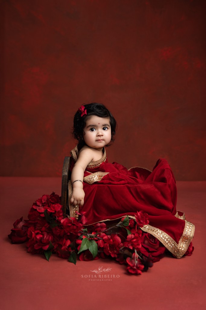 a sweet portrait of a baby girl in traditional Indian attire against a red backdrop