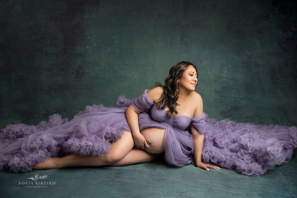 a purple ruffled gown creates a dramatic side laying shot for mom to be, hugging her growing belly with her hands