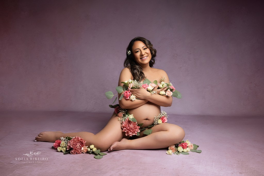 mom is posed against a pink backdrop by a maternity photographer draped in flowers