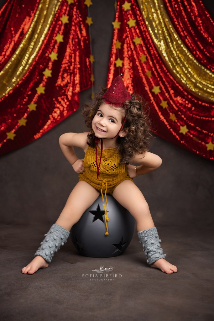 a little girl in a circus hat sits atop a star ball, her expression showing her full personality for a custom vintage birthday session in monmouth county nj