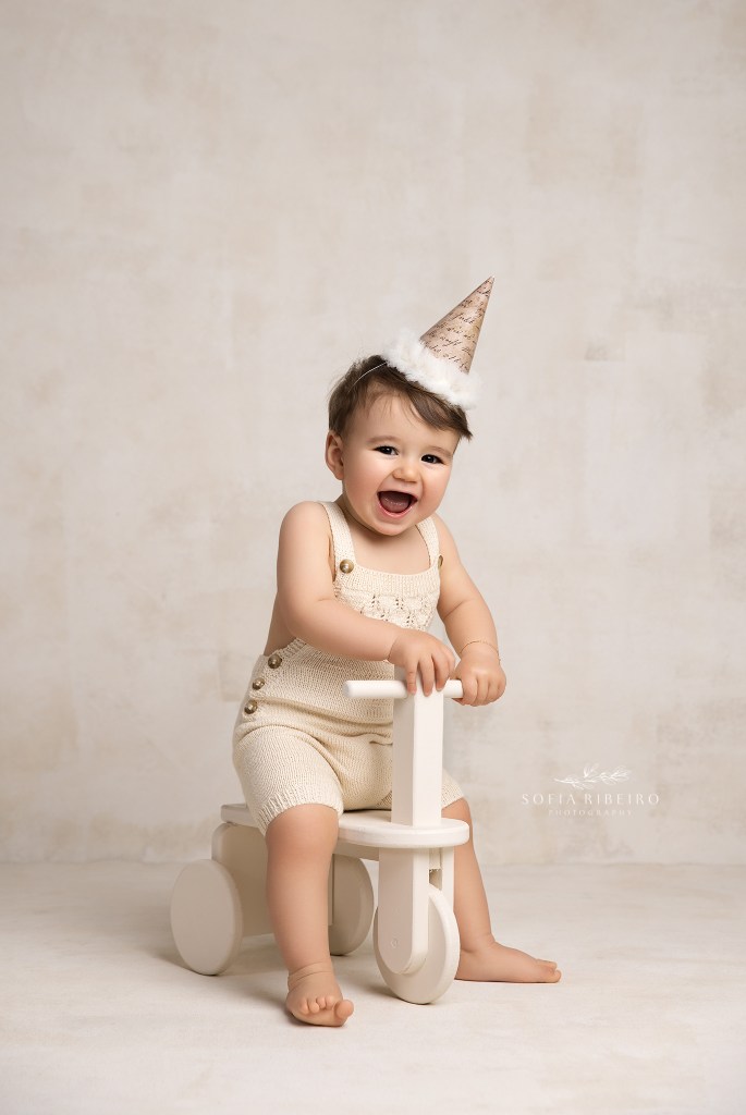 a giggling baby smiles to camera on a wooden tricycle with a fun birthday hat for a portrait session by a monmouth county nj baby photographer