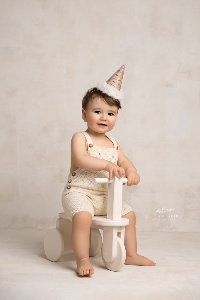 a sweet smiling baby in a cream birthday hat poses for a photo on a wooden tricycle prop