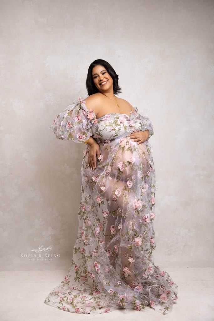 mom poses with hands on belly,showcasing her growing bump in a sheer floral gown