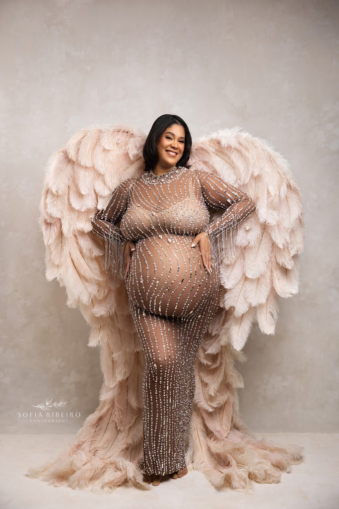 a mom is posed in sparkling dress with soft wings against a beige backdrop by a nj maternity photographer