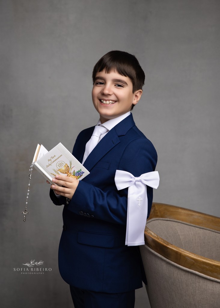 a young boy smiles to camera as he holds his miniature bible and rosary, posing against an armchair for his first communion photos.