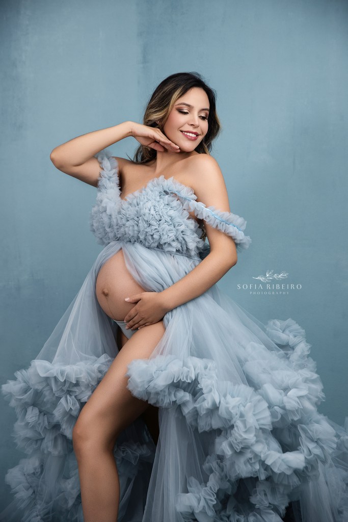 mom strikes a pose cradling her belly with a pale blue tulle dress against a matching backdrop