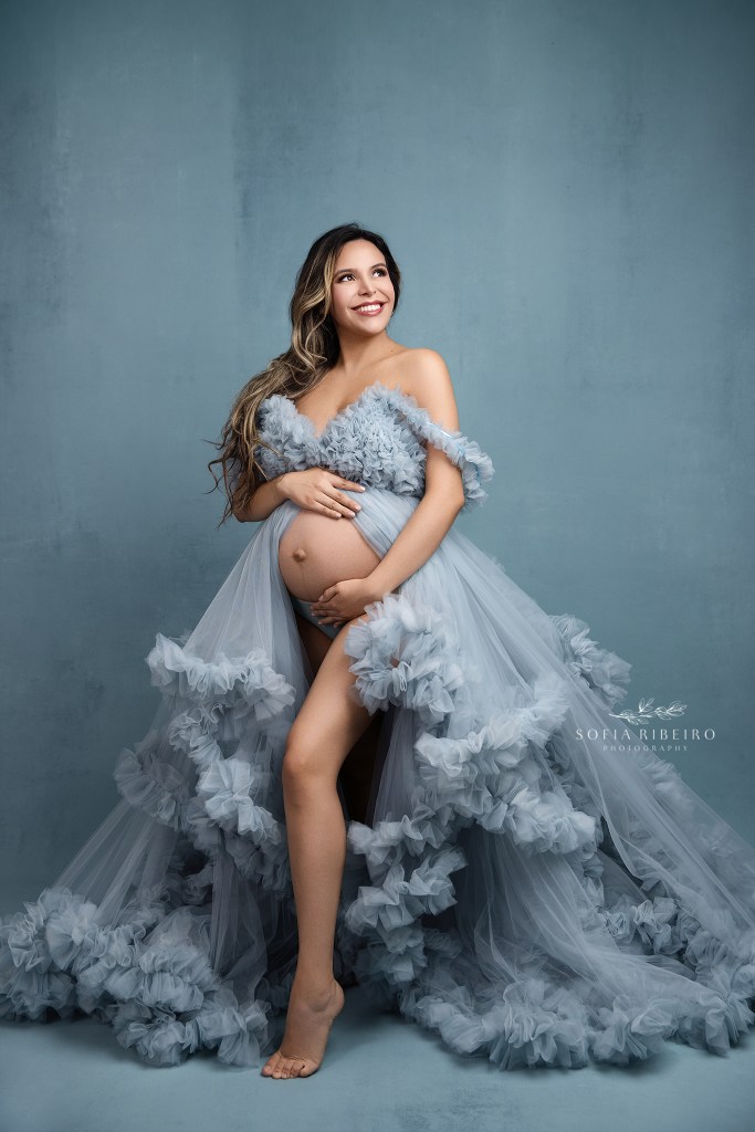 mom strikes a pose cradling her belly with a pale blue tulle dress against a matching backdrop