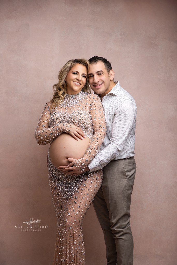 mom and dad to be smile to camera and embrace a sweet moment in celebration of their coming baby
