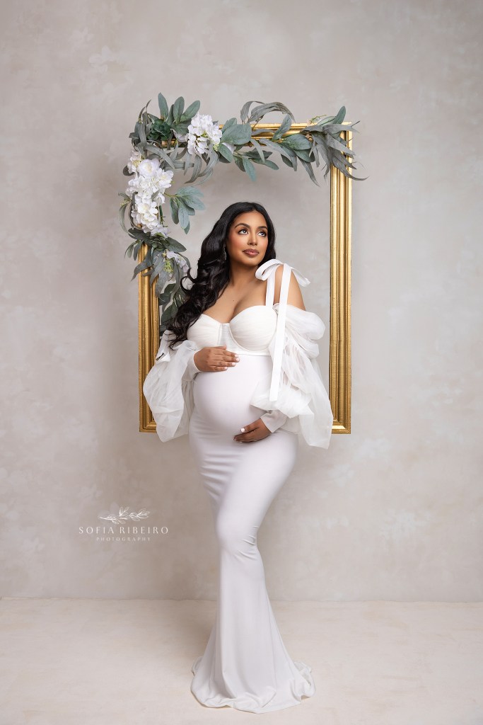 mom to be is beaming in a white belly hugging dress set against a gold floral frame for a maternity portrait in passaic nj