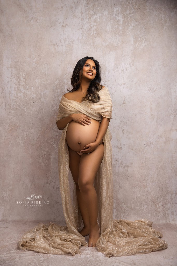a mom to be poses in shimmering gold fabric, smiling and cradling her baby bump.