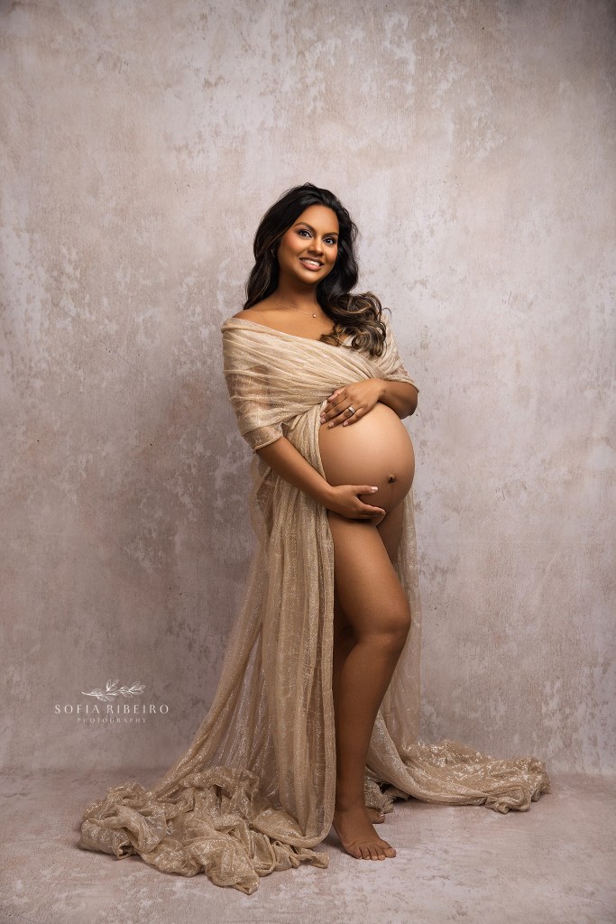 a shimmering gold goddess of a mom to be sparkles in a gold draped fabric against a neutral backdrop