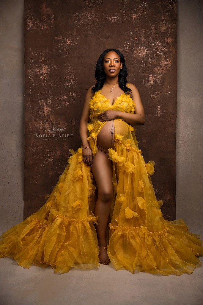 an expectant mom strikes a regal pose in a yellow floral gown against a brown backdrop for maternity portraits in franklin nj