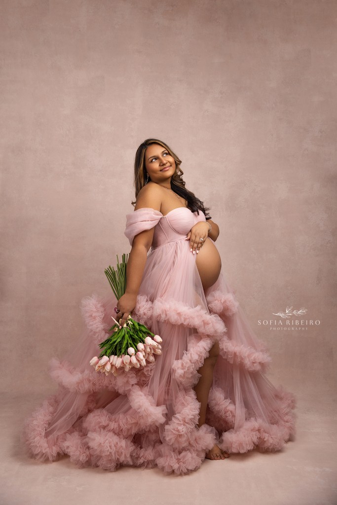 a pink ruffled gown with an open front, captures mom's growing belly while she poses with flowers for a north brunswick nj maternity photographer