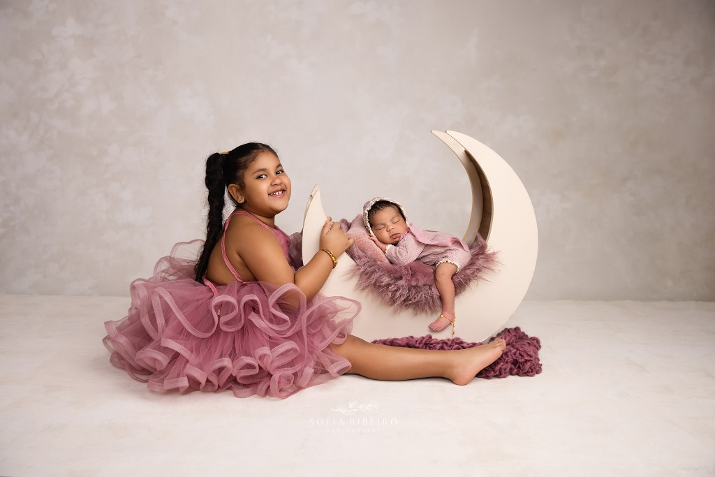 big sister smiles to camera while holding a moon prop with her newborn sister sleeping in it