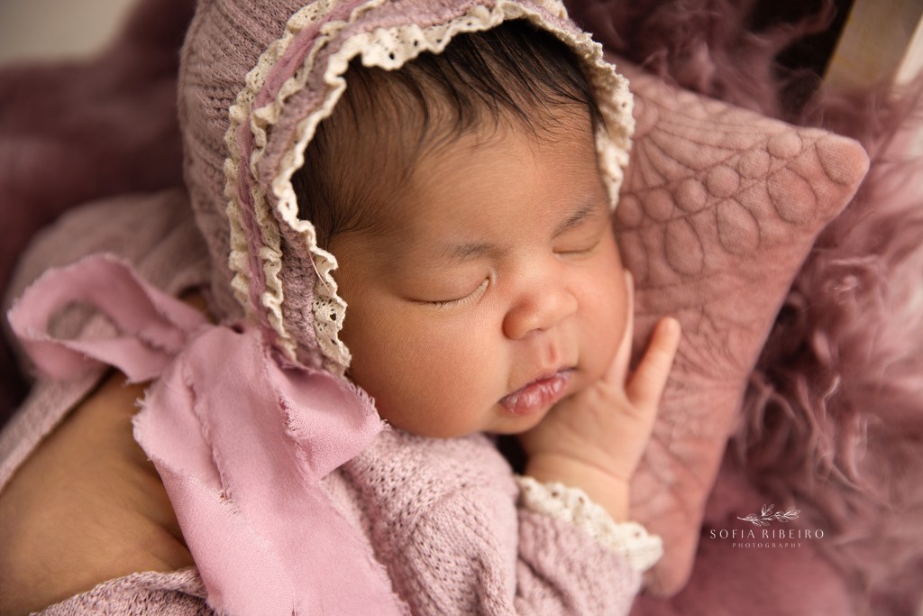 a closeup of a newborn baby girl in a pink bonnet laying on a pillow