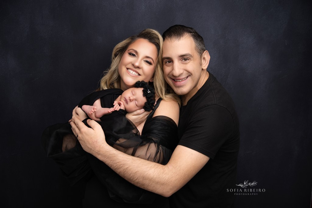 mom and dad smile to camera in all black with their newborn baby girl for a holmdel nj newborn photographer