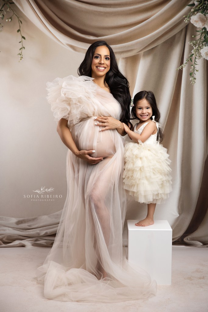 mom and her daughter both cradle her baby bump in matching cream tulle looks against a draped backdrop