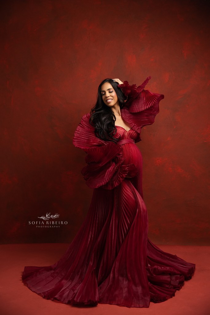 new york city maternity photographer poses mom in an artful read pleated dress showcasing her baby bump