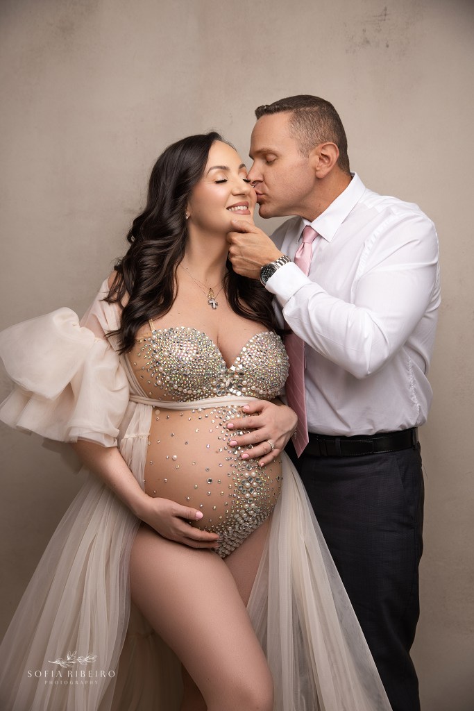 Husband and wife sharing an intimate maternity moment in soft tones of neutral, while mom cradles belly and dad gives her a kiss