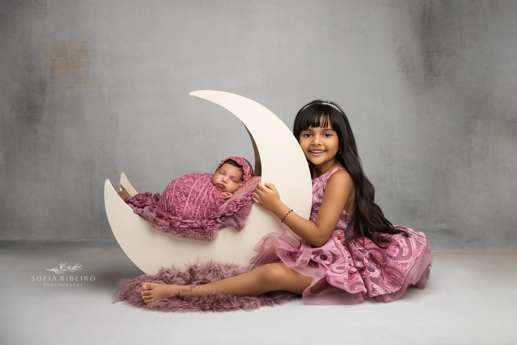 big sister smiles proudly at her baby sister cradled in a wooden moon prop for a portrait in a newborn studio