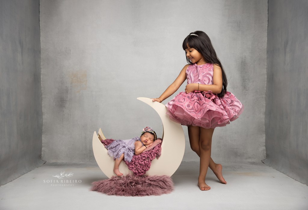 big sister smiles proudly at her baby sister cradled in a wooden moon prop by an east brunswick nj newborn photographer