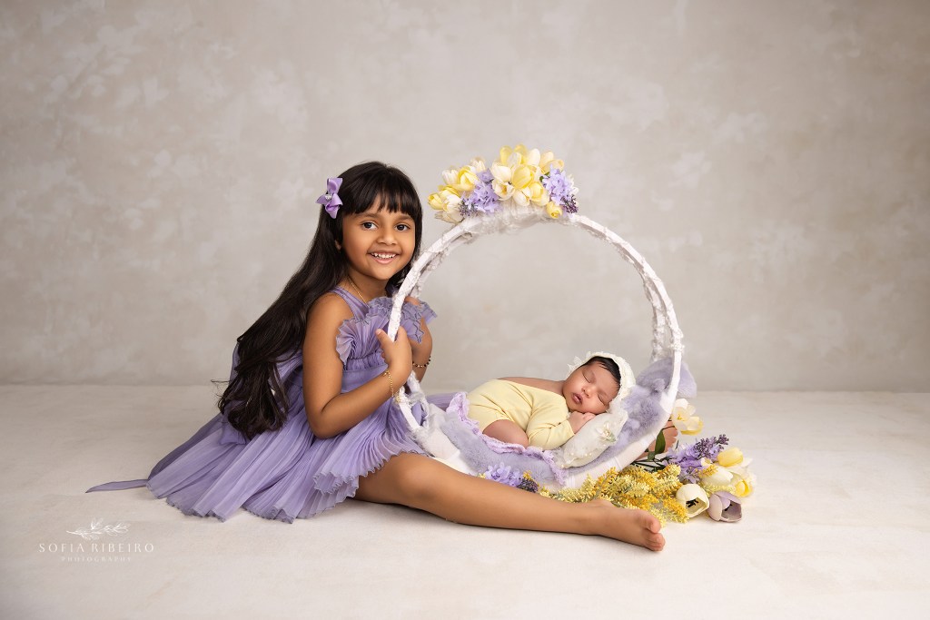 big sister poses proudly with her baby sister in a floral hoop in shades of purple and yellow in an east brunswick nj newborn photographer's studio