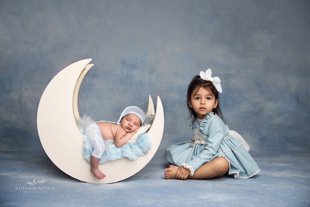Newborn session with big sister posing in a moon prop, captured in Essex County NJ, in matching shades of blue