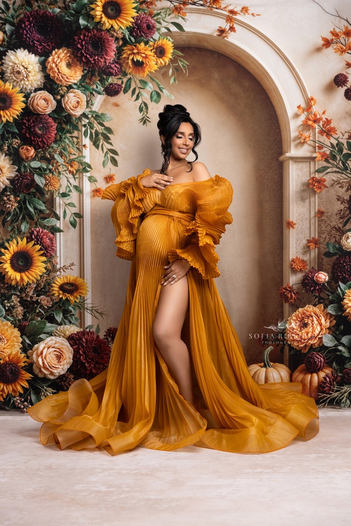 mom to be poses elegantly in a pleated golden gown against a fall floral arch backdrop