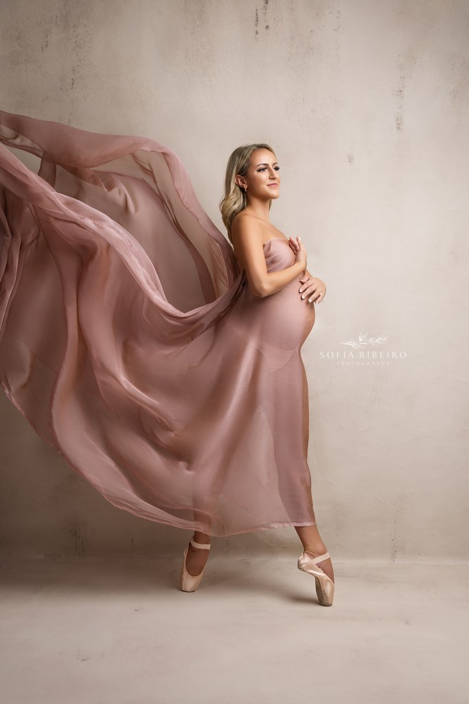 pregnant mom is a ballerina and poses on point shoes and flowing pink fabric for a maternity photographer in ridgewood nj