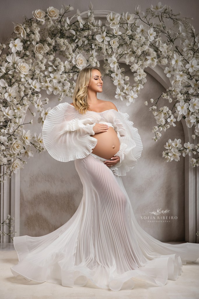a mom exposes her baby bump in a white pleated two piece outfit against a white floral backdrop for photos in ridgewood nj