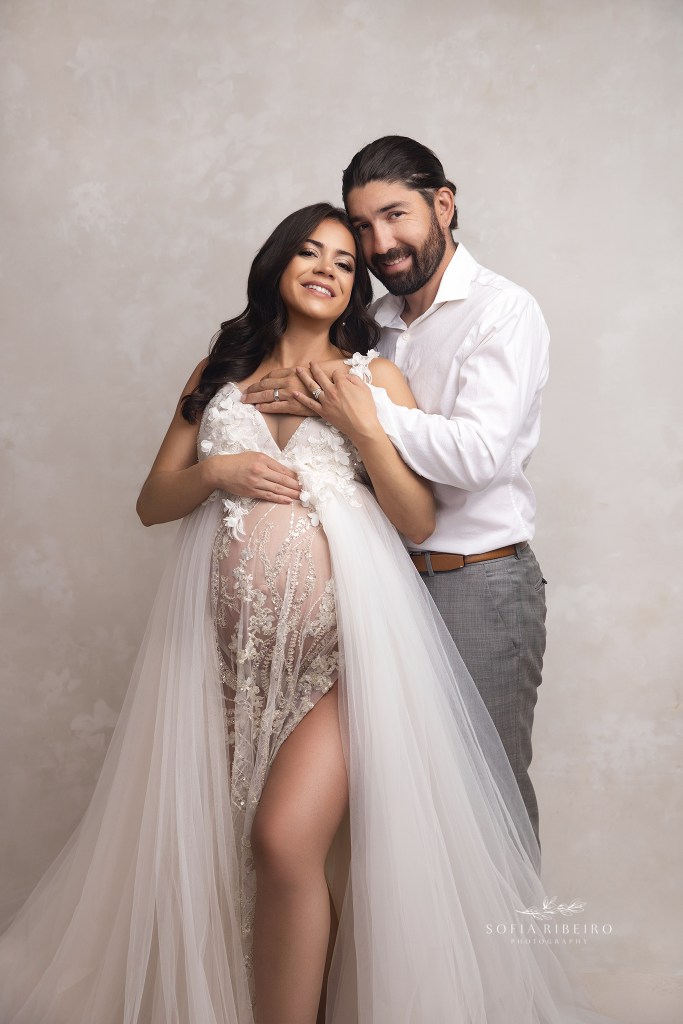 a striking couple poses together in formal attire against a neutral backrop for maternity portraits