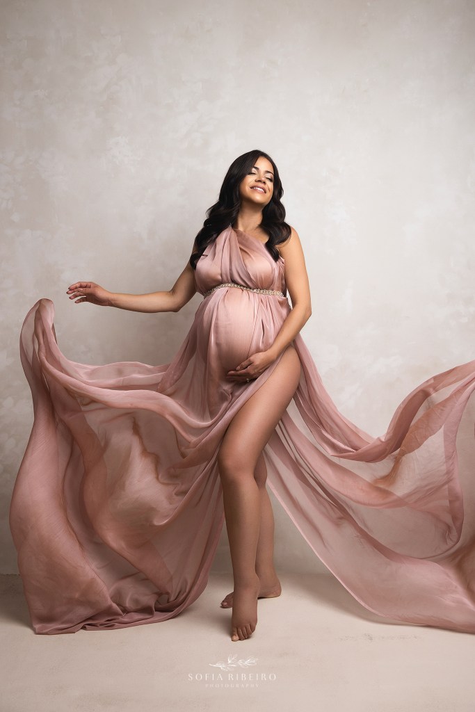 a pregnant mom holds flowing pink fabric, framing her baby bump against a cream backdrop in a maternity studio in cedar grove nj